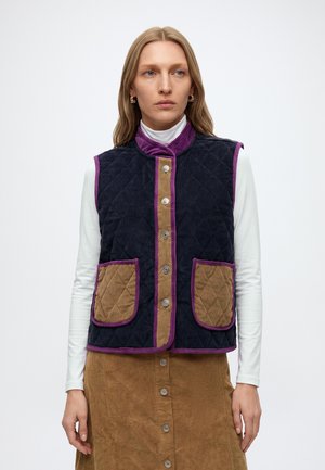 Mulher a usar um colete matelassê azul-marinho com acabamento roxo e bolsos castanhos por cima de uma blusa branca de manga comprida e uma saia castanha com botões, em pé contra um fundo branco.