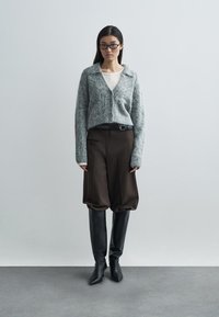 Grijze, zachte cardigan met een kraag, gecombineerd met bruine, oversized broek en zwarte, kniehoge laarzen, voorzien van een riem en een gladde textuur.