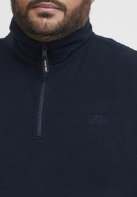 Pullover in felpa blu navy con colletto a mezza zip, caratterizzato da un logo sul petto e una superficie morbida e texture.
