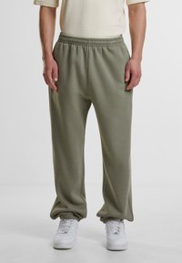 Olivegrønne joggebukser med elastisk midje og mansjetter, laget av mykt materiale. Kombinert med hvite joggesko, sitter de litt løst.