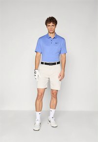 Férfi golf ruházat: világoskék galléros póló, bézs rövidnadrág, fekete öv, fehér zokni és fehér sportcipő. Golfkesztyű a bal kézen.