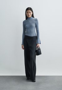 Kék és szürke csíkos garbó, magas derekú fekete csíkos nadrággal párosítva. Egy kis fekete táska a kezében. Sima textúrák.