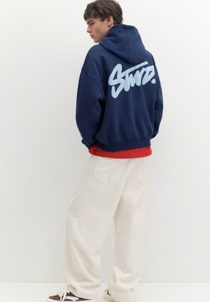 Junger Mann trägt einen dunkelblauen Hoodie mit weißer Schrift auf dem Rücken, rotes Shirt darunter, weite weiße Hose und braune Sneakers mit weißen Schnürsenkeln.