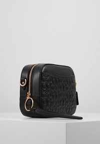 Musta nahkainen olkalaukku, jossa kohokuvioitu kuviointi, kultaiset metalliosat ja vetoketjutarkin. Varustettu irrotettavalla hapsulla ja ryhdikkäällä muodolla.