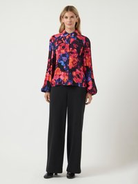 Mulher vestindo uma blusa de manga longa floral brilhante com calças largas pretas e sapatos pretos, de pé contra um fundo claro e simples.