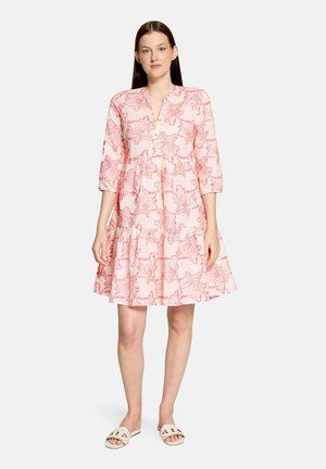 Femme portant une robe rose mi-longue aux motifs floraux rouges et des manches trois-quarts, associée à des sandales blanches à bout ouvert.