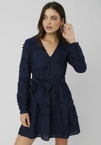 Robe bleu marine à manches longues, décolleté en V, motif floral brodé, taille cintrée avec un lien, et ourlet à volants superposés.