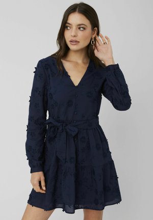 Robe bleu marine à manches longues, décolleté en V, motif floral brodé, taille cintrée avec un lien, et ourlet à volants superposés.