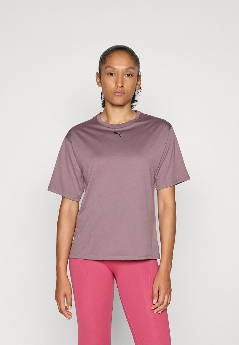 O femeie cu părul strâns într-un coc, îmbrăcată cu un tricou mauve Puma și colanți roz, stând pe un fundal simplu deschis.