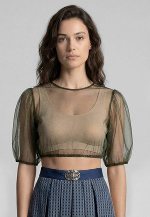 Woman wearing a sheer olive green cropped blouse with puff sleeves over a beige top and a high-waisted blue skirt with a decorative clasp.