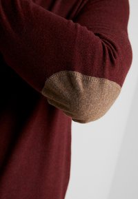 Pull bordeaux avec coudières marron, fabriqué en tissu doux, design ajusté et texture côtelée le long des manches et du col.