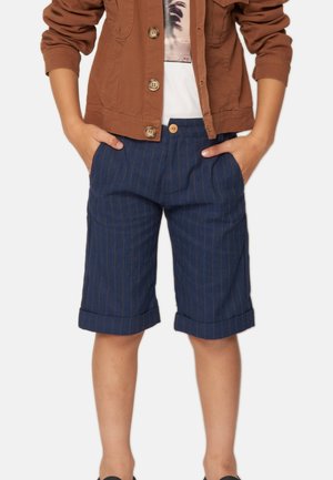 Giacca marrone sopra una camicia bianca, abbinata a pantaloni corti a righe blu navy. I pantaloni corti presentano risvolti e tasche frontali. Scarpe nere visibili.