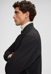 Zwarte gestructureerde sweatshirt met een hoge kraag en witte opstaande kraag. Weergegeven op een persoon in profiel tegen een effen achtergrond.