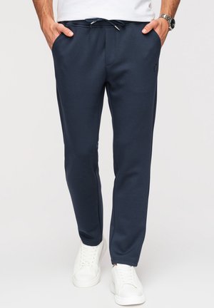 Homme portant un pantalon de jogging bleu marine avec taille à cordon, des baskets blanches et une chemise blanche, les mains dans les poches, debout devant un fond uni.