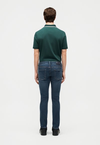 Άνδρας που στέκεται με την πλάτη στραμμένη, φοράει σκούρο πράσινο κοντομάνικο πόλο μπλουζάκι, μπλε στενά τζιν παντελόνια και μαύρα παπούτσια, σε λευκό φόντο.