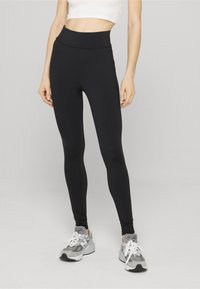 Svarte leggings med høy midje, laget av et elastisk materiale. Modellen kombinerer med grå sneakers og viser glatt tekstur og tettsittende design.