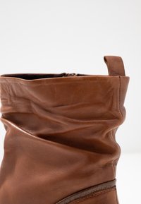 Brauner, weicher Lederstiefel mit einem lässigen Schaft und einer Schlaufe an der Rückseite, vor einem schlichten weißen Hintergrund.