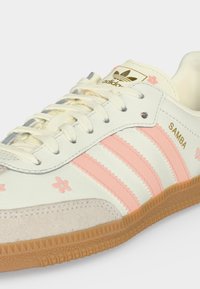 Λευκό αθλητικό Adidas Samba με ανοιχτορόζ ρίγες, χρυσό λογότυπο, λευκοκρεμ κορδόνια, καφέ σόλα και μικρές ανοιχτορόζ λουλουδένιες λεπτομέρειες στο επάνω μέρος.