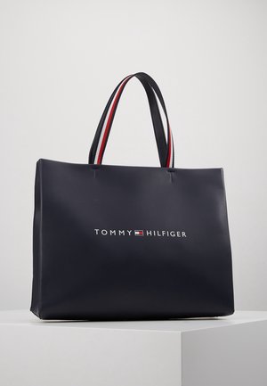 Mørkeblå Tommy Hilfiger tote-taske med røde, hvide og sorte dobbelte håndtag, vist på en hvid overflade mod en ensfarvet baggrund.