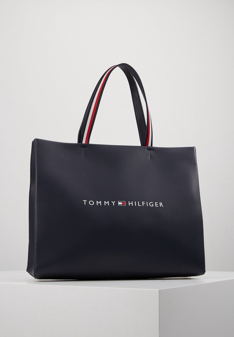 Sac cabas bleu marine Tommy Hilfiger avec doubles poignées rouges, blanches et noires, présenté sur une surface blanche avec un fond uni.
