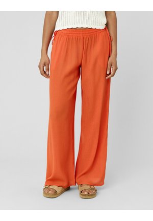 Femme portant un pantalon ample orange vif à taille élastique et des sandales beige, debout devant un fond uni.