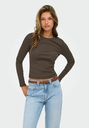 Femme aux longs cheveux ondulés portant une chemise à manches longues brun foncé, un jean bleu clair et une ceinture brune tressée, debout avec une main sur la hanche.