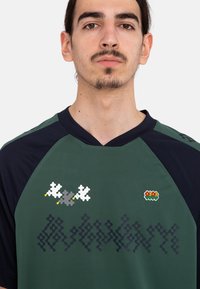 Jersey de manga corta en verde oscuro y azul marino que presenta un patrón geométrico, flores bordadas y dos acentos gráficos coloridos en la parte frontal.