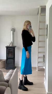 Top preto de mangas longas, saia midi azul, botins pretos. Chão de madeira clara, paredes brancas, chaminé de metal e um sofá ao fundo.