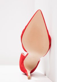 Escarpin en daim rouge à talon haut et bout pointu, avec la semelle tournée vers l'avant, reposant contre un coin de mur et de sol blancs.