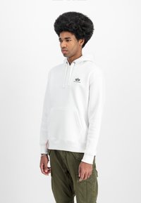 White hoodie with a front pocket, drawstring hood, and embroidered logo on the chest. Paired with olive green cargo pants.