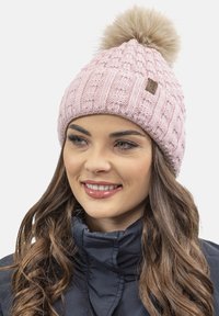 Gorro de lana rosa con un patrón texturizado, adornado con un pom-pom de piel sintética de color marrón claro. Presenta una pequeña etiqueta marrón en el lateral.
