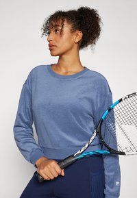 Blauer Baumwollpullover mit gerippten Bündchen, kombiniert mit marinefarbenen Leggings. Das Model hält einen Tennisschläger mit einem schwarz-blauen Design.