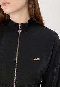 Zwarte jas met hoge kraag en een ritssluiting aan de voorkant, met een logo-accent in metallic goud, gemaakt van gestructureerde stof.