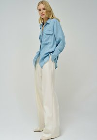 Camisa de denim de gran tamaño en azul claro con dos bolsillos frontales, combinada con pantalones de pierna ancha en color crema y zapatos planos beige, sobre un fondo neutro.