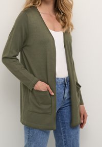 Olivově zelený svetr na knoflíky z měkké látky, s výstřihem do V, otevřeným předním dílem a dvěma předními kapsami; nošený přes bílý top a modré džíny.