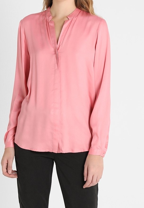 Femme portant un chemisier rose à manches longues avec un petit col en V et un pantalon noir, debout devant un fond clair uni.