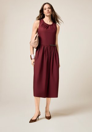 Bordeaux mouwloze jurk met geplooide halslijn en elastische taille, gecombineerd met een beige clutch en schoenen met luipaardprint. Soepele stoftextuur.