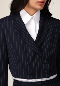 Blazer doppiopetto a righe navy con camicia bianca sotto. Presenta una vestibilità su misura e cuciture in contrasto di colore argento.