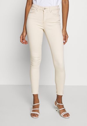 Kvinde iført beige high-waist skinny jeans med frynsete kanter og hvide hesteløbssandaler med hæl, stående mod en ensfarvet baggrund.