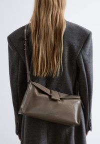 Braune Leder-Umhängetasche mit Klappenverschluss und verstellbarem Schulterriemen, gehalten gegen einen grauen oversized Blazer. Glatte Textur, minimalistisches Design.