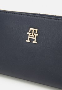 Navy Lederportemonnaie mit goldener Logo-Akzentuierung. Die Oberfläche hat eine glatte Textur und einen Reißverschluss entlang der oberen Kante.