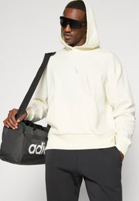 Hoodie branca texturizada com capuz, logotipo preto no peito, combinada com calças de fato pretas e uma mala de desporto preta com um logotipo branco.