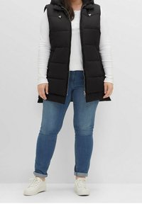 Schwarze, gepolsterte Weste mit Kapuze, Reißverschluss und silbernen Details, getragen über einem weißen Langarmoberteil und blauen Jeans. Weiße Sneakers sind ebenfalls enthalten.