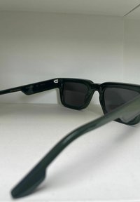 Zwarte zonnebril met een glanzende afwerking, vierkante lenzen en een dik montuur. Inclusief branding op de poot en een label voor UV-bescherming van categorie 3.