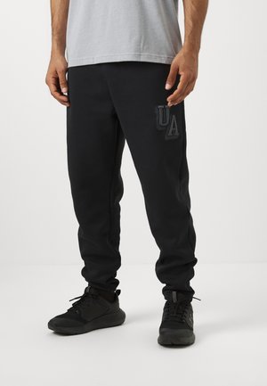 Zwarte sweatpants van zacht materiaal, met een rubberen "UA"-logo op de linker bovenbeen. Getailleerde fit met elastische boorden, gecombineerd met zwarte sneakers.