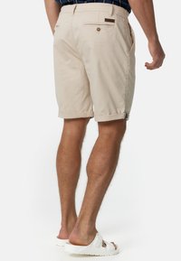 Man draagt beige shorts met opgerolde zoom en witte slide sandalen, staat met zijn rug naar de camera gericht tegen een effen achtergrond.