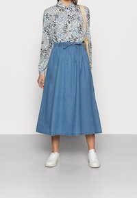 Gonna maxi in denim azzurro con vita regolabile, abbinata a una blusa a maniche lunghe con fantasia. Sneakers bianche completano l'outfit.