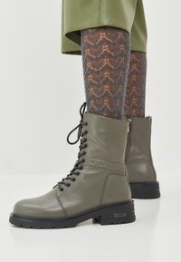 Gevlochten combatlaarzen in olijfgroen leer met een gemêleerd patroon, chunky zwarte zool en decoratieve hardwareaccenten.