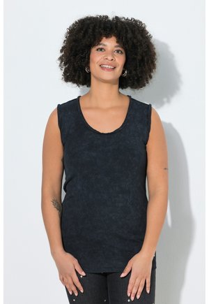 Uśmiechnięta kobieta o kręconych włosach, ubrana w czarny top bez rękawów i ciemne spodnie, na jasnym, jednolitym tle, z rękami swobodnie opuszczonymi wzdłuż ciała.