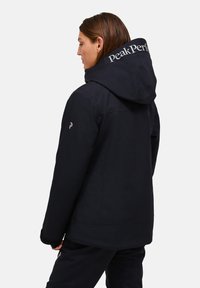 Giacca nera con cappuccio, dotata di colletto alto. Logo "Peak Performance" in bianco sul retro. Materiale liscio e impermeabile.